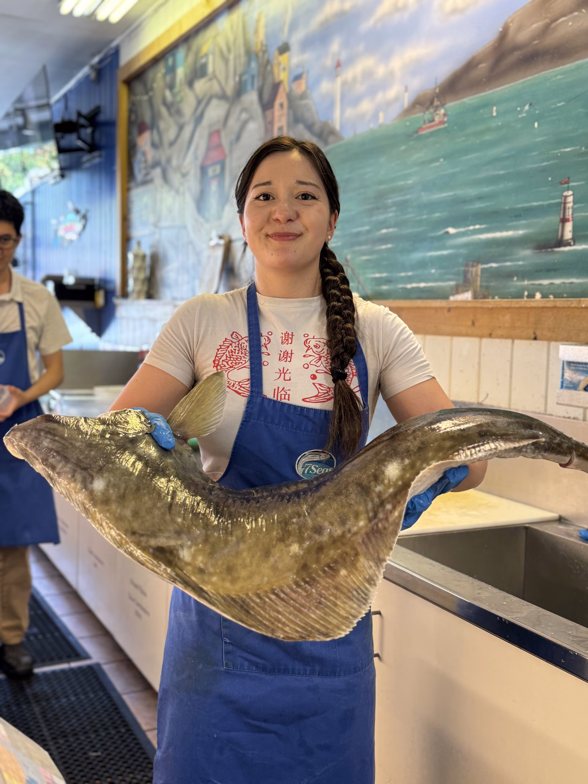 A 7 Seas fishmonger holding a whole Pacific halibut in front of the counter